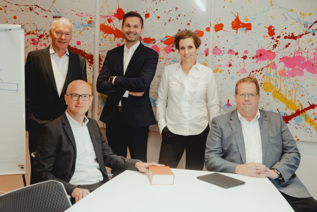 Das Team steht zusammen an einem Tisch vor einem Bild